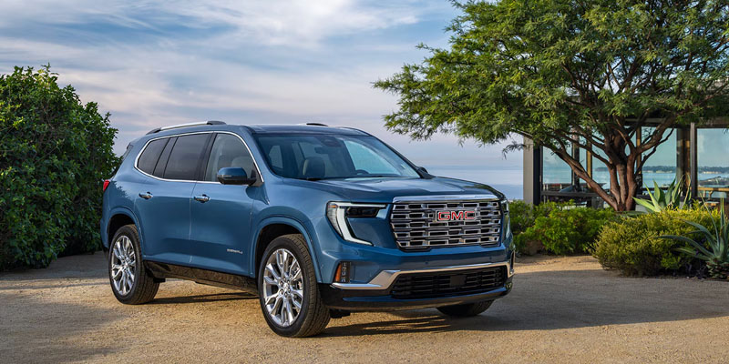 A sky-blue 2026 GMC Acadia shines under the sky as it is parked beside a lake with green trees and bushes on the shore - a lovely, harmonious sight to see.