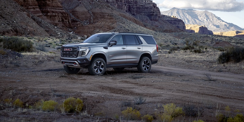 A shining grey 2026 GMC Yukon SUV is parked on a bushy, flat area at the base of a desert mountain.