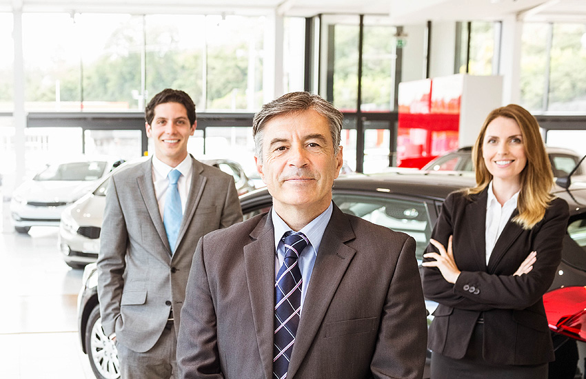 2 grey-suited GMC salesmen and a long-haired blonde saleswoman with folded hands smile at us as they stand in front of the GMC vehicles for sale in the sunlit many-windowed salesroom.